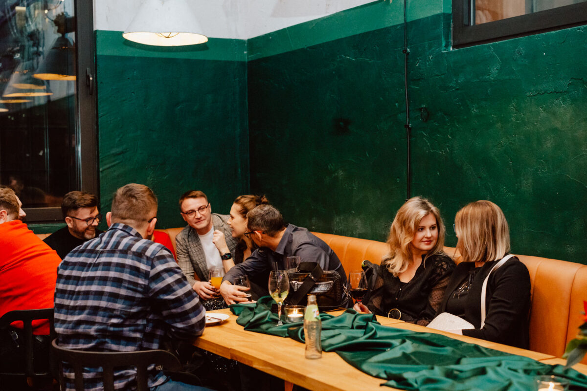 wigilia firmowa spoldzielnia koneser fotograf na impreze 038 Grupa ludzi siedzi razem przy długim drewnianym stole w przytulnej restauracji z zielonymi ścianami, rozmawiając i śmiejąc się, podczas gdy na stole stoją napoje i świece.