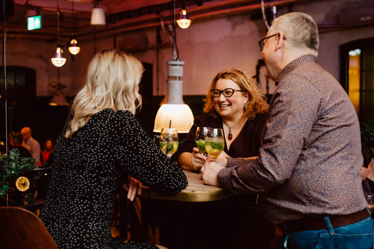 wigilia firmowa spoldzielnia koneser fotograf na impreze 039 Trzy dorosłe osoby siedzą przy małym stoliku w słabo oświetlonym barze, uśmiechając się i rozmawiając, trzymając koktajle z limonką i miętą, otoczone ciepłymi wiszącymi światłami i industrialnym wystrojem.