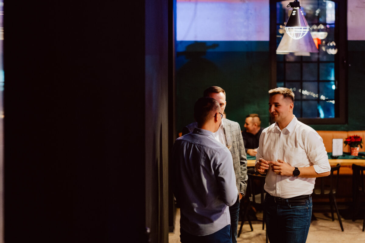 wigilia firmowa spoldzielnia koneser fotograf na impreze 040 Trzech mężczyzn stoi i rozmawia w słabo oświetlonej restauracji lub barze. Dwie kobiety siedzą przy stoliku w tle, a przez okno widać światła sznurkowe.