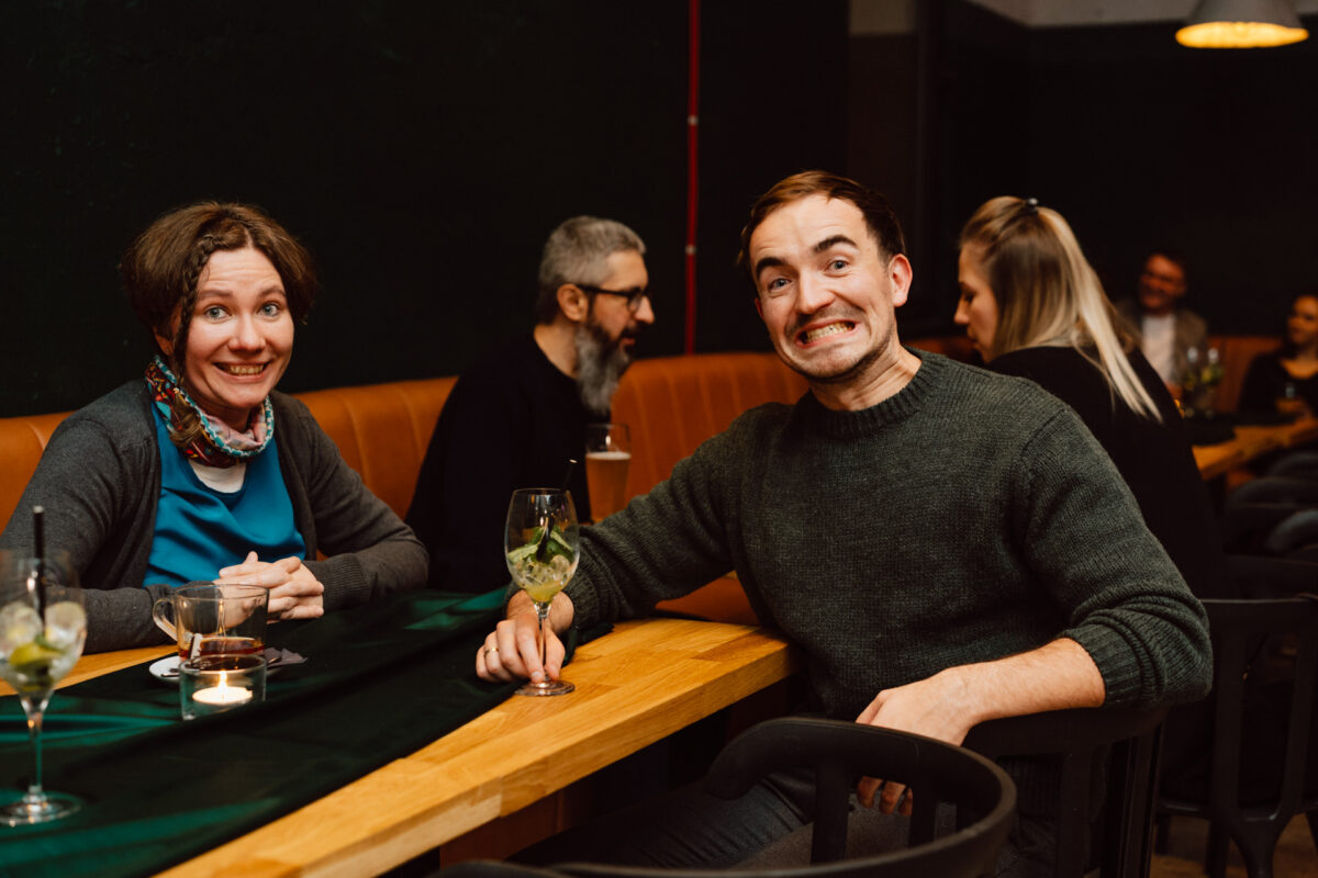 wigilia firmowa spoldzielnia koneser fotograf na impreze 041 Dwie osoby siedzą przy drewnianym stole w przytulnym barze lub restauracji, uśmiechając się do kamery. Inni klienci są widoczni w tle, a na stole stoją napoje. Atmosfera wydaje się ciepła i zrelaksowana.