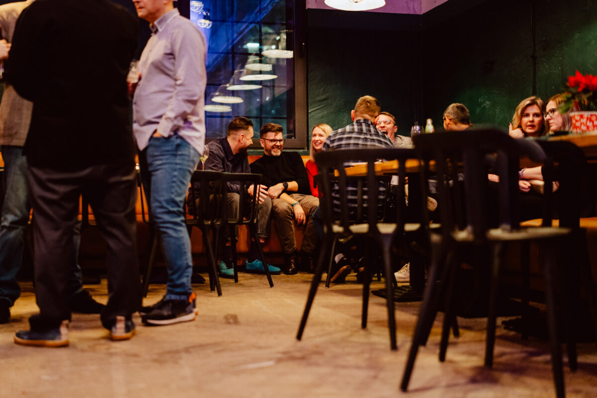 wigilia firmowa spoldzielnia koneser fotograf na impreze 042 Grupa ludzi siedzi i stoi przy stolikach w słabo oświetlonej restauracji lub barze, rozmawiając i śmiejąc się. Niektórzy siedzą, podczas gdy inni stoją, tworząc ciepłą i żywą atmosferę.