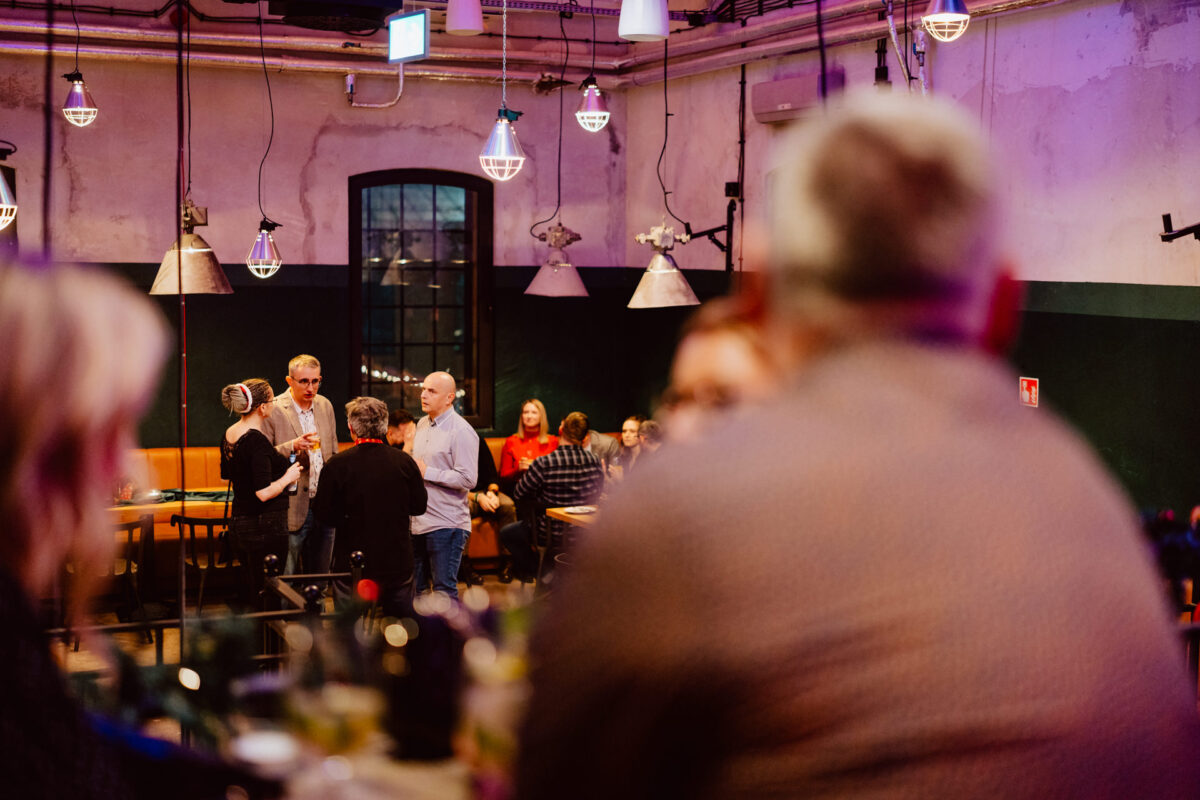 wigilia firmowa spoldzielnia koneser fotograf na impreze 043 Ludzie zgromadzeni w ciepło oświetlonym, nowoczesnym barze lub restauracji, rozmawiający w małych grupach. Ostrość skupia się na grupie stojącej w pobliżu okna, podczas gdy inni siedzą lub stoją na delikatnie rozmytym pierwszym planie.