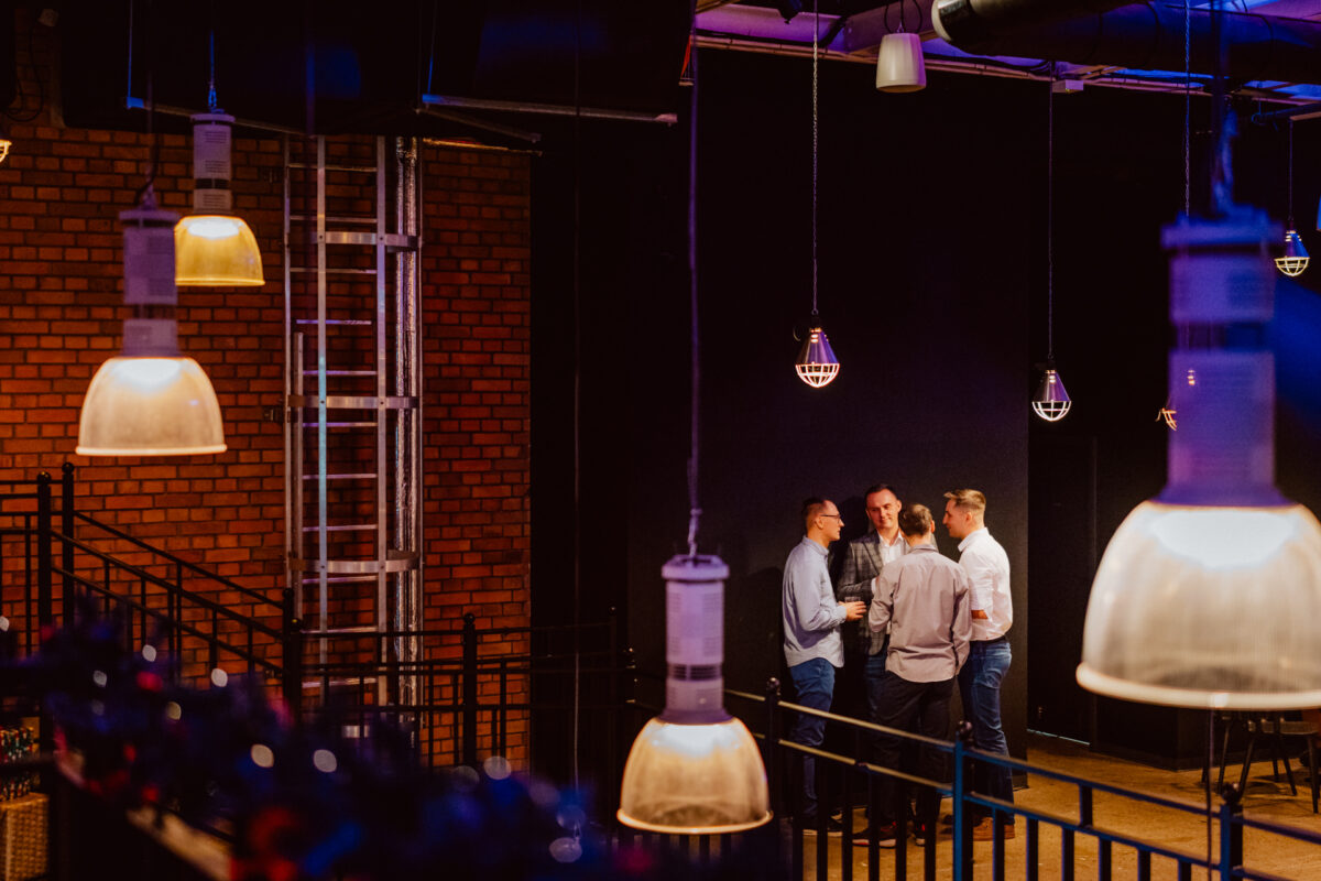 wigilia firmowa spoldzielnia koneser fotograf na impreze 044 Grupa pięciu osób rozmawia przy ciepłym świetle wiszących lamp w industrialnym pomieszczeniu z ceglanymi ścianami i metalową drabiną.