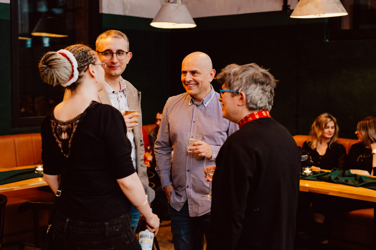 wigilia firmowa spoldzielnia koneser fotograf na impreze 045 Cztery osoby stoją w grupie, uśmiechają się i rozmawiają, trzymając drinki w ciepło oświetlonej restauracji. W tle inni goście siedzą i rozmawiają przy stolikach.