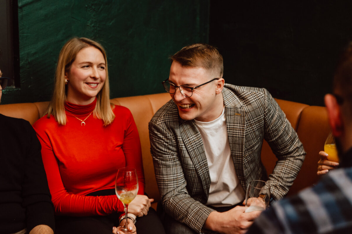 wigilia firmowa spoldzielnia koneser fotograf na impreze 046 Kobieta w czerwonym golfie i mężczyzna w marynarce w kratę siedzą na opalonej kanapie, uśmiechając się i śmiejąc razem w przytulnym wnętrzu, każde z nich trzyma drinka.