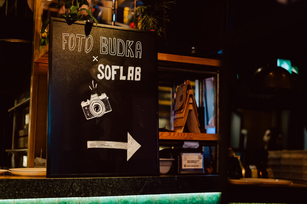 wigilia firmowa spoldzielnia koneser fotograf na impreze 047 Tablica z napisem "FOTO BUDKA x SOFLAB" z rysunkiem aparatu i białą strzałką skierowaną w prawo, umieszczona na drewnianej ladzie z roślinami i półkami w słabo oświetlonym pomieszczeniu.