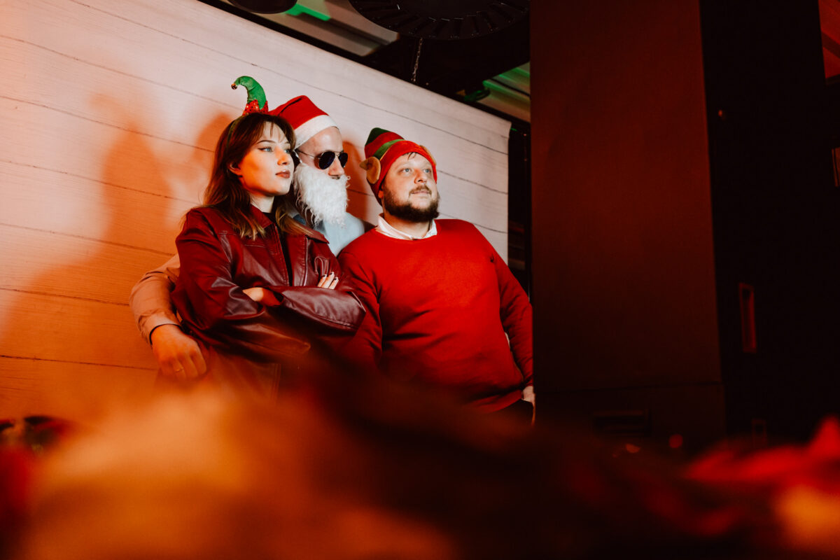 wigilia firmowa spoldzielnia koneser fotograf na impreze 048 Trzy dorosłe osoby pozują w pomieszczeniu na tle jasnej drewnianej ściany, ubrane w świąteczne stroje. Jedna osoba nosi czapkę Mikołaja i okulary przeciwsłoneczne, inna nosi opaski o tematyce świątecznej, a wszyscy mają wesołą, zrelaksowaną postawę.