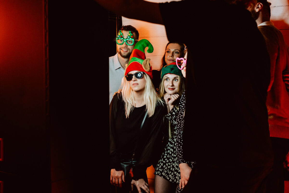 wigilia firmowa spoldzielnia koneser fotograf na impreze 049 Cztery osoby pozują do świątecznego zdjęcia w zabawnych akcesoriach, takich jak świąteczne czapki, okulary przeciwsłoneczne i nowatorskie okulary. Uśmiechają się i stoją blisko siebie na ciepło oświetlonym tle.