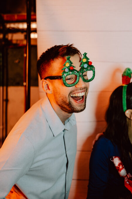 wigilia firmowa spoldzielnia koneser fotograf na impreze 050 Mężczyzna w nowatorskich okularach w kształcie choinki i jasnoniebieskiej koszuli śmieje się radośnie. Kobieta obok niego, częściowo poza kadrem, nosi świąteczną opaskę w kolorach czerwonym i zielonym. Tło jest proste i jasne.