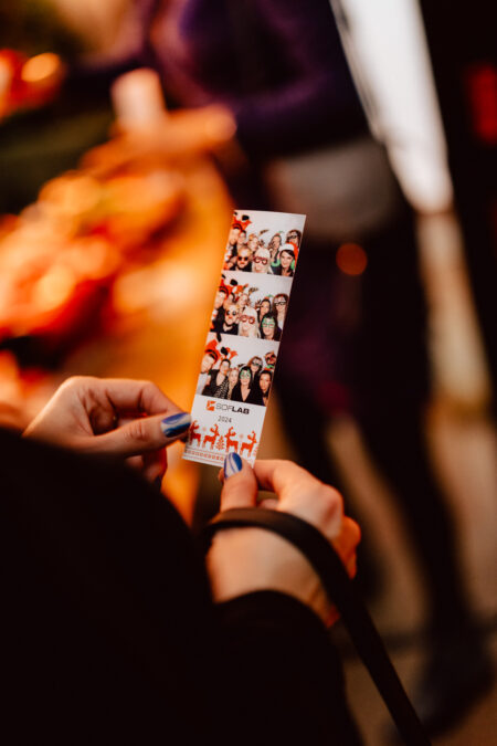 wigilia firmowa spoldzielnia koneser fotograf na impreze 051 Osoba z pomalowanymi na niebiesko paznokciami trzyma pasek zdjęć przedstawiający kilka uśmiechniętych osób ubranych w świąteczne akcesoria. Tło jest rozmyte, ukazując niewyraźnych ludzi i ciepłe światła.