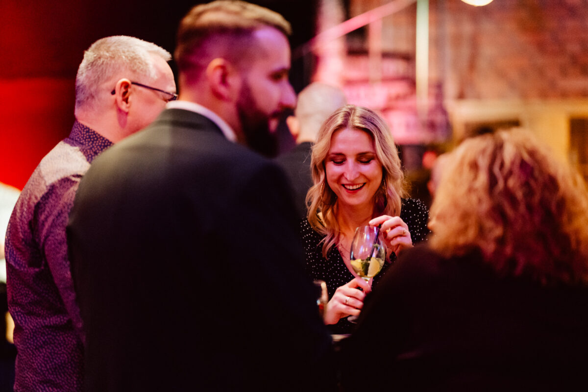 wigilia firmowa spoldzielnia koneser fotograf na impreze 054 Grupa czterech osób spotyka się w słabo oświetlonym pomieszczeniu. Uśmiechnięta kobieta o blond włosach trzyma kieliszek wina, zaangażowana w rozmowę z innymi przy stole. Tło jest ciepło oświetlone w odcieniach czerwieni i żółci.