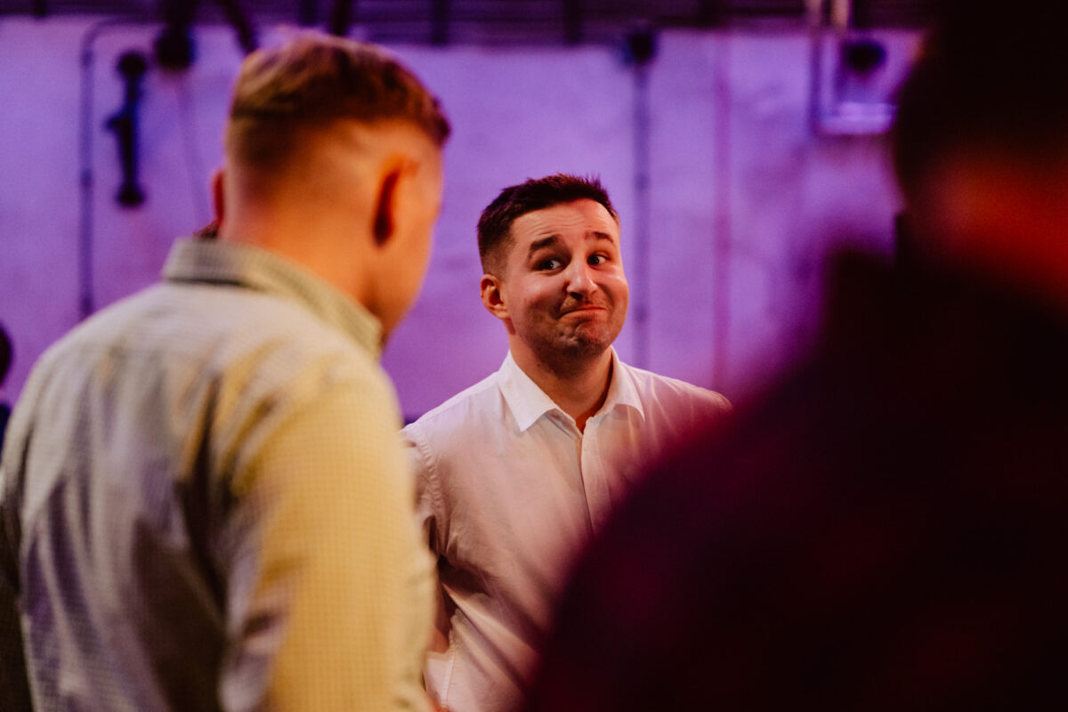 wigilia firmowa spoldzielnia koneser fotograf na impreze 055 Mężczyzna w białej koszuli stoi w pomieszczeniu z lekkim uśmiechem, patrząc na kogoś przed sobą. Dwie inne osoby są częściowo widoczne, zwrócone w jego stronę. Tło zawiera fioletowe oświetlenie i rozmyte obiekty.