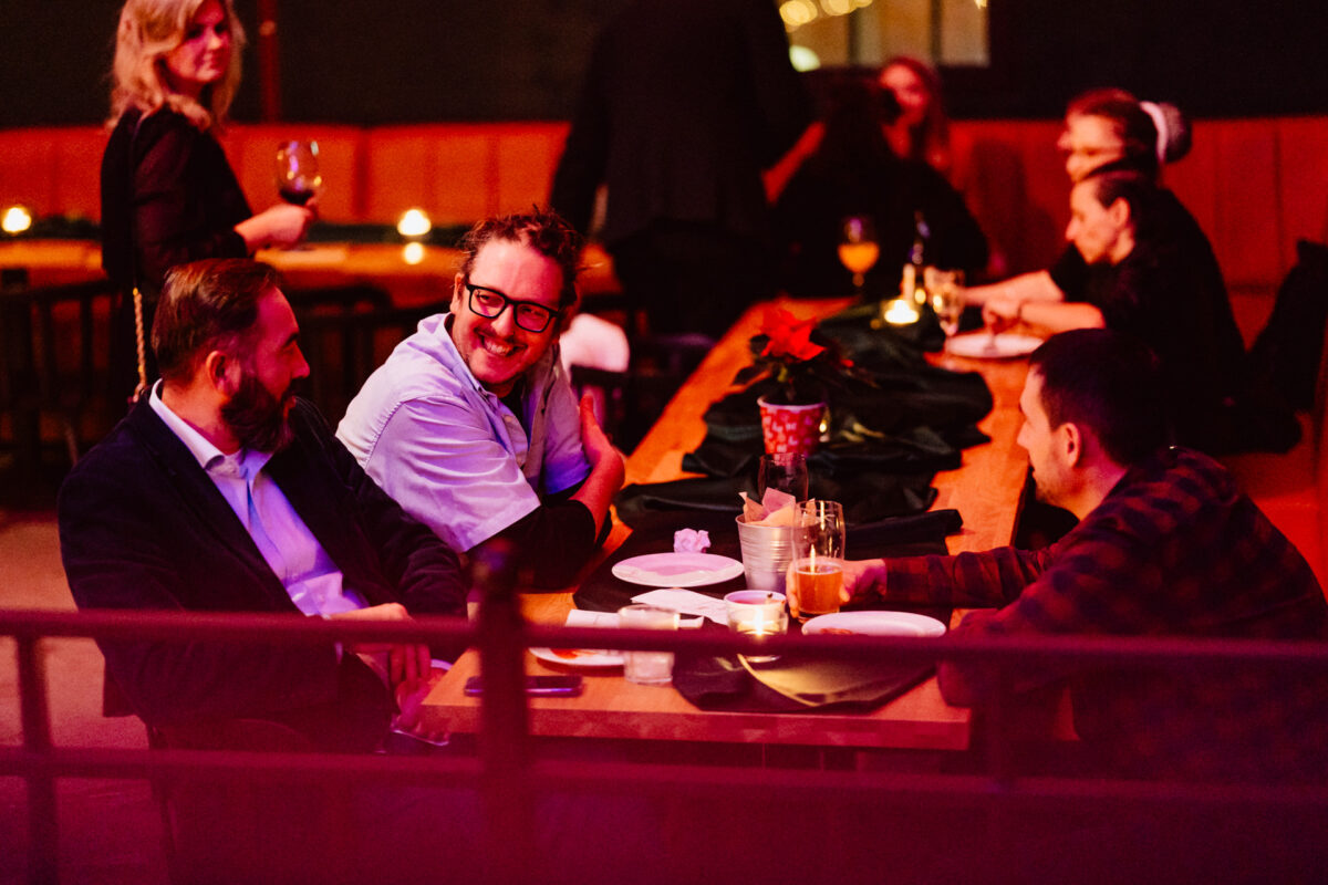 wigilia firmowa spoldzielnia koneser fotograf na impreze 056 Grupa ludzi siedzi wokół drewnianego stołu w słabo oświetlonym barze lub restauracji, rozmawiając i uśmiechając się, z napojami i talerzami przed sobą. Ciepłe, nastrojowe oświetlenie tworzy przytulną atmosferę.