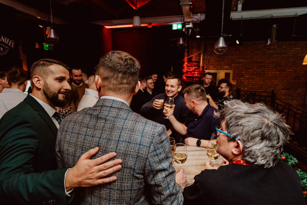 wigilia firmowa spoldzielnia koneser fotograf na impreze 057 Grupa ludzi rozmawiających towarzysko i brzęczących kieliszkami przy drewnianym stole na imprezie w pomieszczeniu, z świątecznym oświetleniem i odsłoniętymi ceglanymi ścianami w tle.