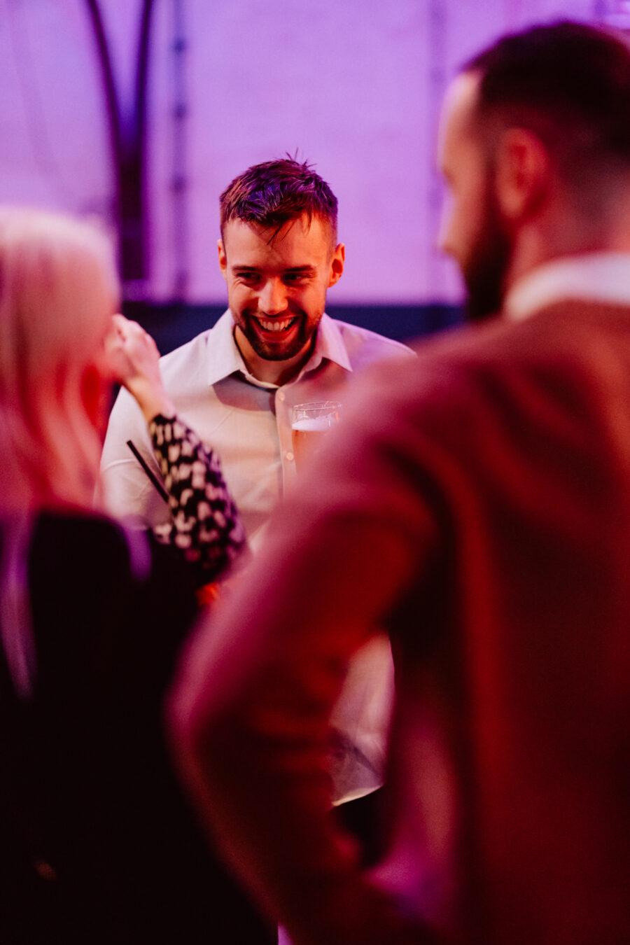 wigilia firmowa spoldzielnia koneser fotograf na impreze 059 Mężczyzna w białej koszuli uśmiecha się i śmieje podczas rozmowy z dwiema innymi osobami na imprezie w pomieszczeniu z ciepłym, różowawym oświetleniem. Scena wydaje się żywa i towarzyska.