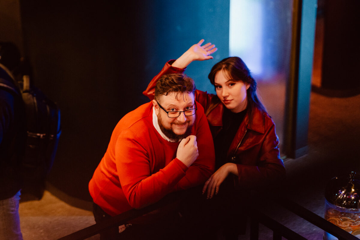 wigilia firmowa spoldzielnia koneser fotograf na impreze 060 Dwie osoby w czerwonych kurtkach stoją w pomieszczeniu przy ciepłym oświetleniu. Jedna osoba uśmiecha się do kamery, trzymając rękę na brodzie, podczas gdy druga stoi obok, podnosząc rękę żartobliwie i patrząc w kamerę.