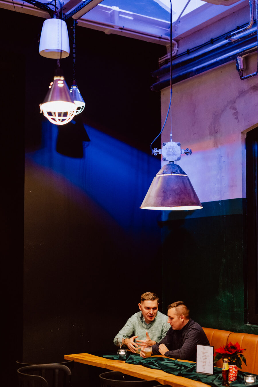 wigilia firmowa spoldzielnia koneser fotograf na impreze 061 Dwóch mężczyzn siedzi przy stoliku w słabo oświetlonej restauracji i prowadzi rozmowę. Trzy duże wiszące lampy nad nimi rzucają ciepły i dramatyczny blask na scenę.