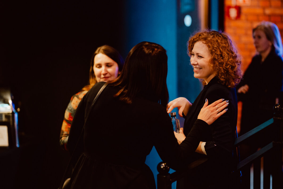 wigilia firmowa spoldzielnia koneser fotograf na impreze 063 Trzy kobiety rozmawiają i uśmiechają się podczas imprezy w pomieszczeniu. Jedna kobieta z kręconymi rudymi włosami śmieje się, podczas gdy inna dotyka jej ramienia. Czwarta kobieta stoi w tle przy ceglanej ścianie. Atmosfera wydaje się przyjazna i ciepła.