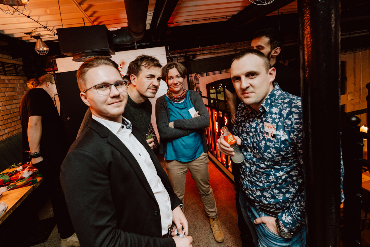 wigilia firmowa spoldzielnia koneser fotograf na impreze 064 Grupa pięciu osób stoi blisko siebie na imprezie towarzyskiej w pomieszczeniu, swobodnie ubrana i uśmiechnięta do kamery. W tle widoczne jest ciepłe oświetlenie i industrialny wystrój.