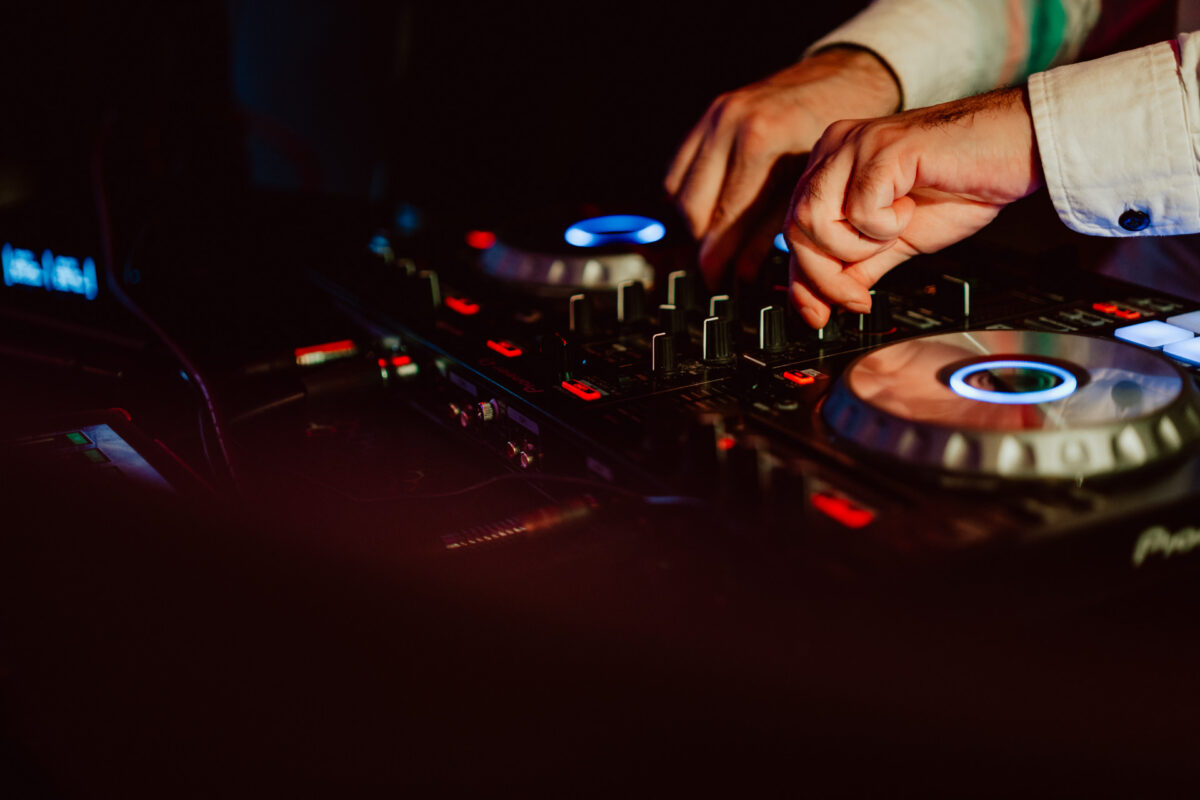 wigilia firmowa spoldzielnia koneser fotograf na impreze 065 Zbliżenie rąk DJ-a regulującego pokrętła i elementy sterujące na profesjonalnym mikserze DJ-skim, z kolorowymi światłami oświetlającymi sprzęt w słabo oświetlonym otoczeniu.