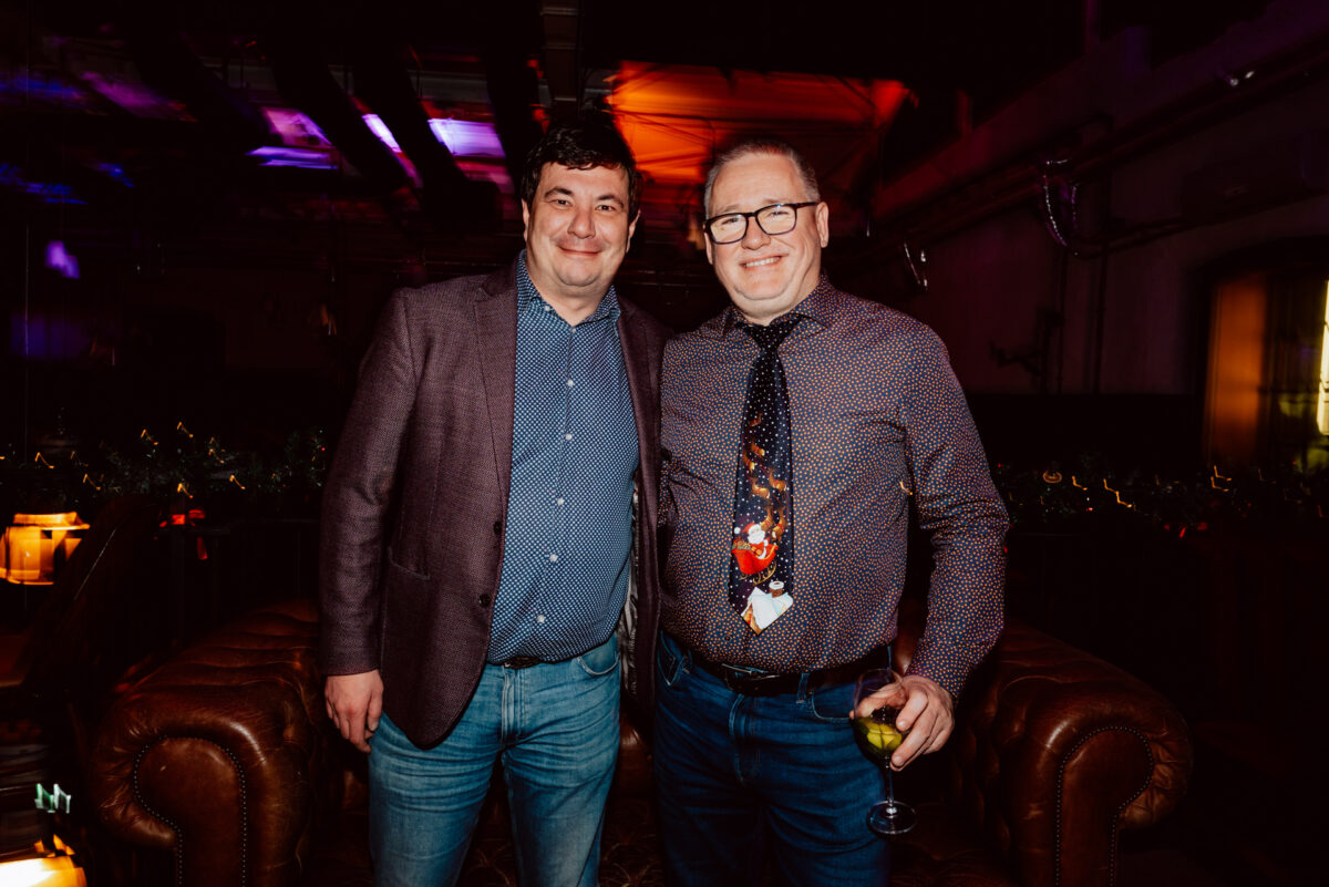 wigilia firmowa spoldzielnia koneser fotograf na impreze 066 Dwóch uśmiechniętych i stojących blisko siebie mężczyzn, ubranych w luźne kurtki i koszule, pozuje do zdjęcia w pomieszczeniu na brązowych skórzanych sofach. Jeden z mężczyzn trzyma drinka; w tle widoczne są świąteczne światła i dekoracje świąteczne.