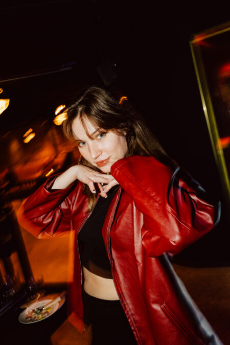 wigilia firmowa spoldzielnia koneser fotograf na impreze 067 Kobieta ubrana w czerwoną skórzaną kurtkę i czarny crop top pozuje z rękami przy brodzie, lekko się uśmiechając, w słabo oświetlonym wnętrzu z ciepłymi światłami w tle.