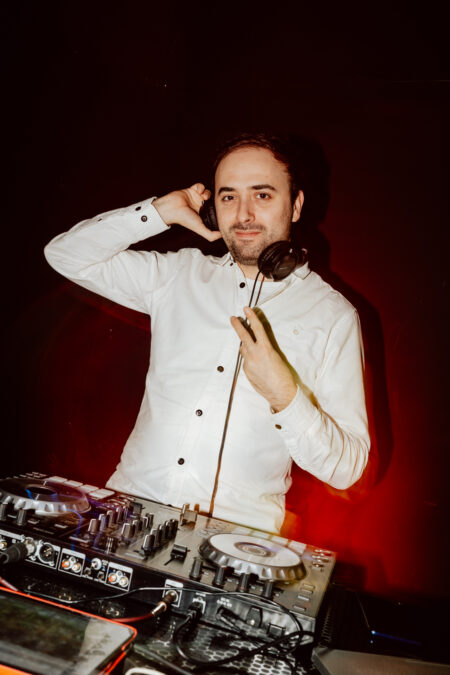 wigilia firmowa spoldzielnia koneser fotograf na impreze 068 DJ w białej koszuli stoi za konsolą mikserską, trzymając słuchawki przy jednym uchu i uśmiechając się do kamery na ciemnym tle z czerwonym oświetleniem.