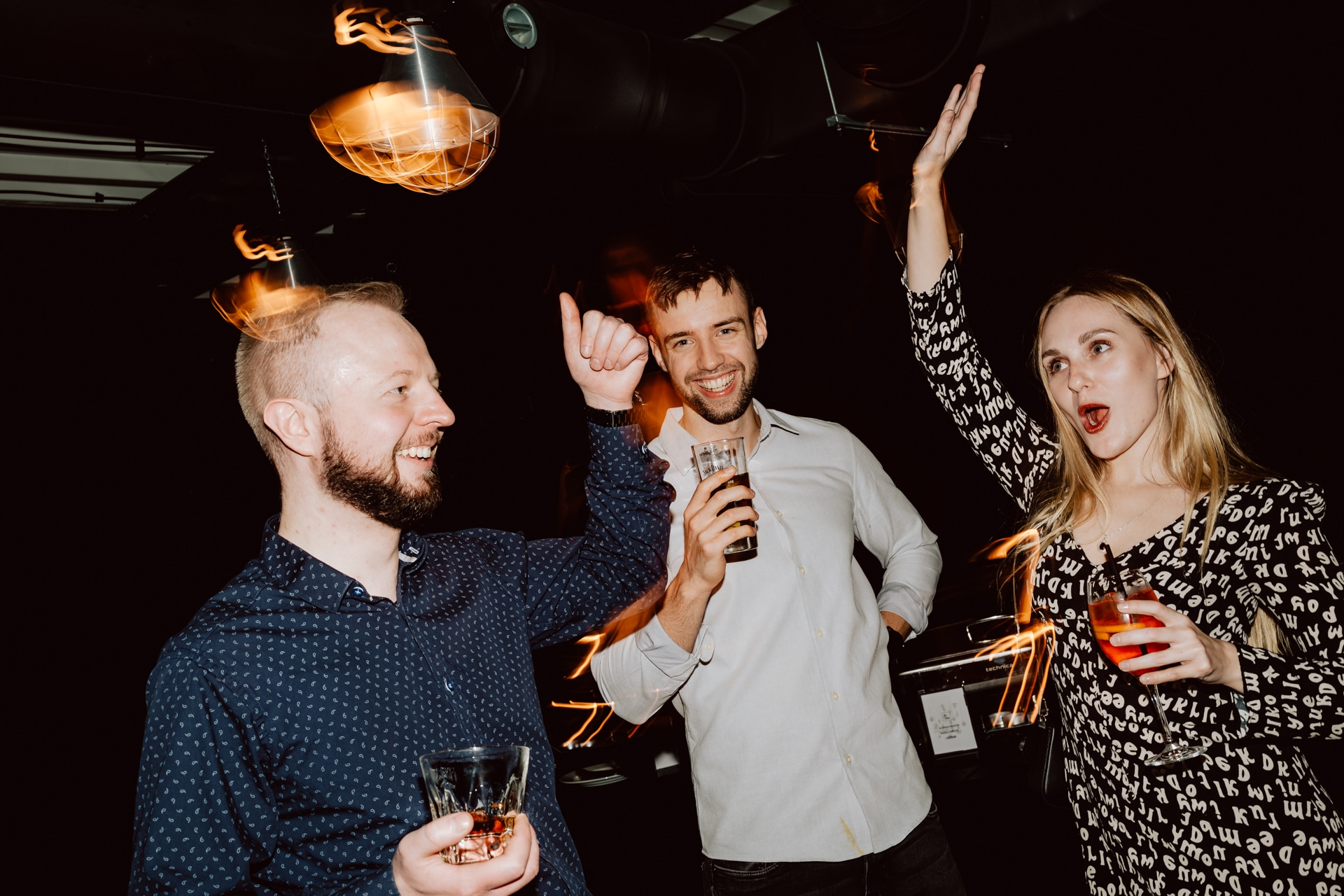 Słabo oświetlony bar z dużym, ozdobnym żyrandolem na pierwszym planie. Na półkach za barem stoją różne butelki z alkoholem. Barman stoi za ladą, a jedna osoba siedzi przed barem.