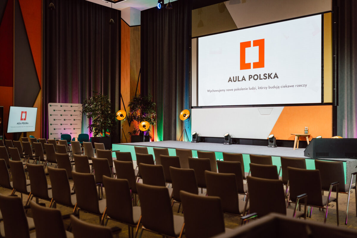 zdjecia eventowe aula polska bnp paribas warszawa 001 Nowoczesna sala konferencyjna z rzędami pustych krzeseł naprzeciwko sceny, dużym ekranem wyświetlającym logo Aula Polska i reflektorami oświetlającymi obszar sceny. Całość wygląda na gotową do prezentacji lub wydarzenia.