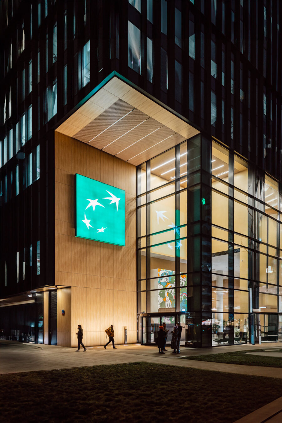 zdjecia eventowe aula polska bnp paribas warszawa 002 Ludzie spacerują nocą przed nowoczesnym, przeszklonym budynkiem. Nad wejściem widnieje duży, podświetlany zielony szyld z białymi sylwetkami ptaków. Wnętrze jest jasno oświetlone i zawiera kolorowe dzieła sztuki.