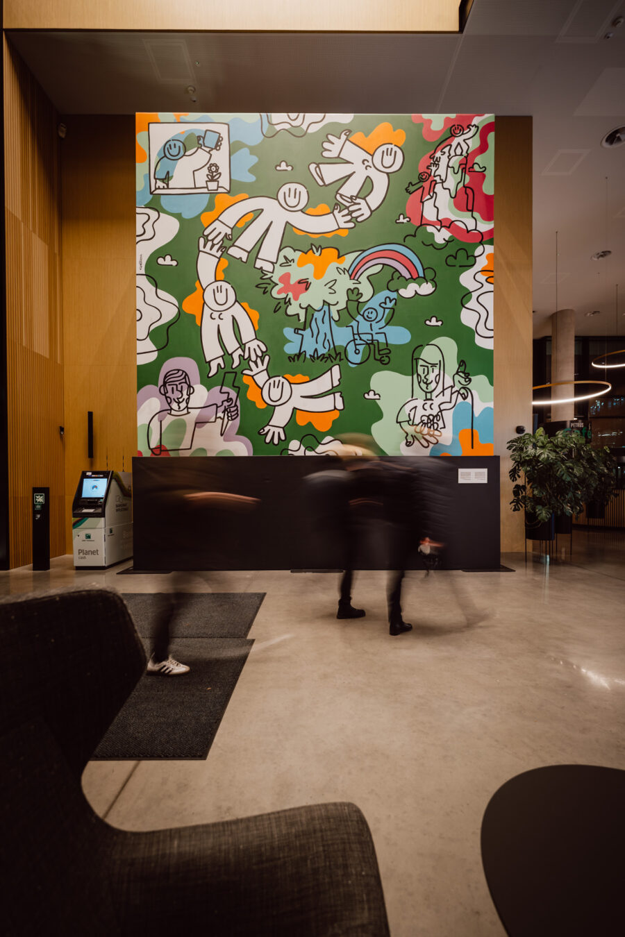 zdjecia eventowe aula polska bnp paribas warszawa 003 Kolorowy mural z wesołymi, kreskówkowymi postaciami, tęczą i chmurami zdobi dużą ścianę w nowoczesnym holu budynku, podczas gdy rozmazani ludzie przechodzą obok po wypolerowanej podłodze.