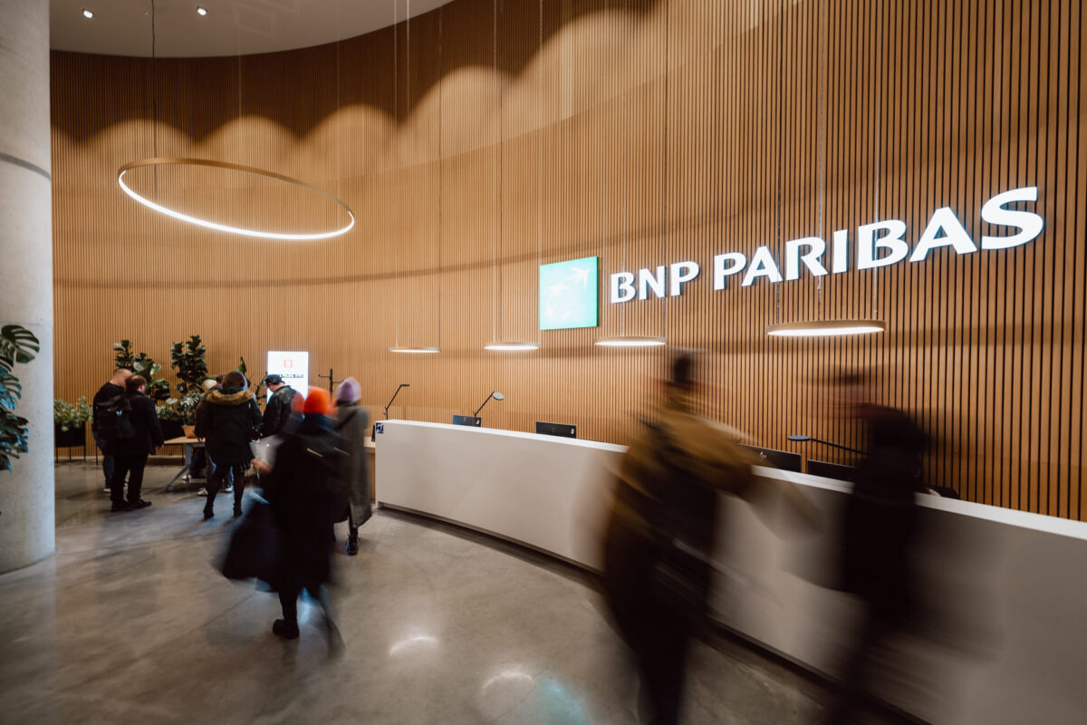 zdjecia eventowe aula polska bnp paribas warszawa 004 Ludzie przechodzą przez nowoczesne, przestronne lobby biura BNP Paribas z zakrzywioną drewnianą ścianą, podświetlanym znakiem, okrągłą oprawą oświetleniową, roślinami i białą recepcją.