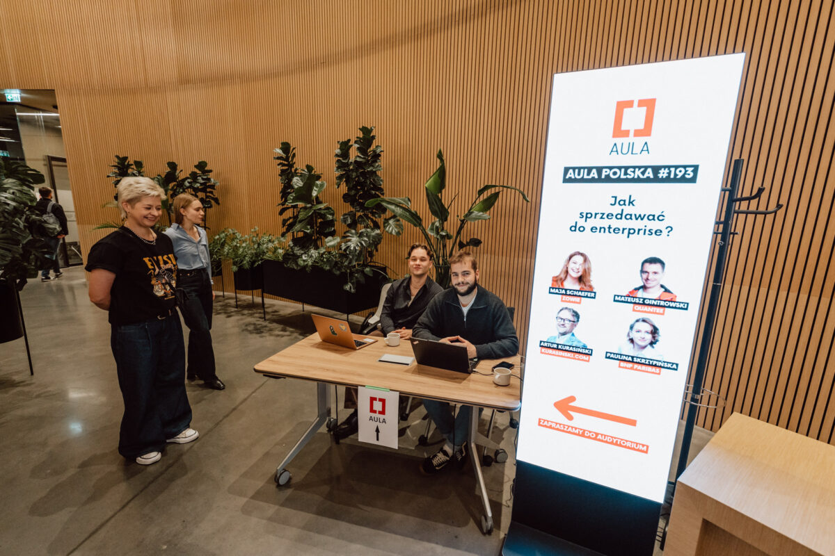 zdjecia eventowe aula polska bnp paribas warszawa 005 Trzy osoby siedzą przy biurku rejestracyjnym z laptopami w nowoczesnym holu, a jedna kobieta stoi obok. Obok nich znajduje się wysoki cyfrowy znak wyświetlający informacje o polskim wydarzeniu o nazwie "Aula Polska #193".