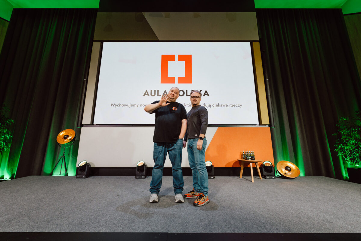 zdjecia eventowe aula polska bnp paribas warszawa 006 Dwie osoby stoją na scenie przed dużym ekranem wyświetlającym logo "Aula Polska". Jedna osoba żartobliwie gestykuluje ręką, podczas gdy druga stoi z rękami w kieszeniach. Scena jest oświetlona zielonymi i pomarańczowymi światłami.