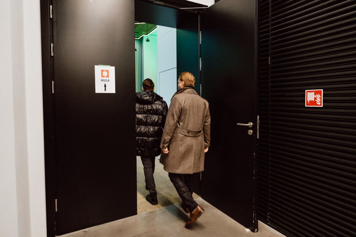 zdjecia eventowe aula polska bnp paribas warszawa 007 Dwie osoby w płaszczach przechodzą przez duże otwarte drzwi do korytarza. Znak na drzwiach z napisem "AULA" i strzałką wskazuje wnętrze. Na ścianie po prawej stronie widoczny jest znak bezpieczeństwa przeciwpożarowego.