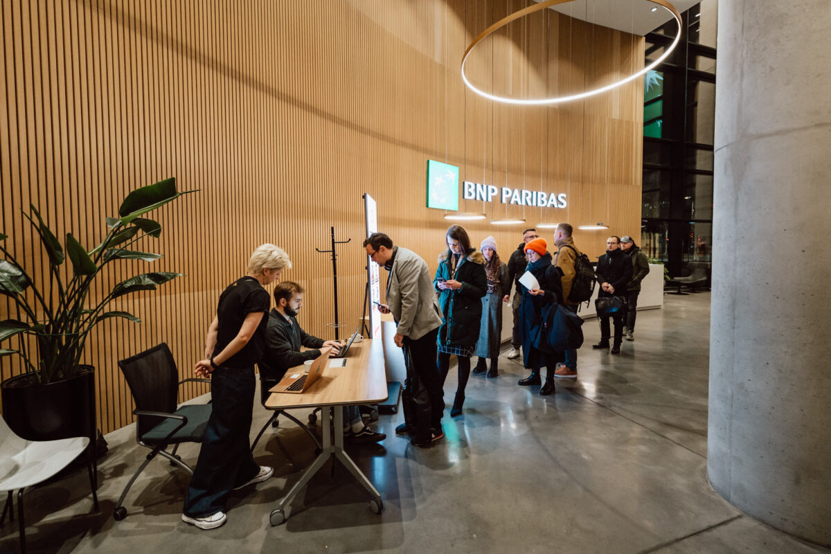 zdjecia eventowe aula polska bnp paribas warszawa 008 Ludzie stoją w kolejce do rejestracji w nowoczesnym, dobrze oświetlonym biurze BNP Paribas, a personel im pomaga. W tle widoczne są drewniane ściany, okrągła oprawa oświetleniowa i rośliny doniczkowe.