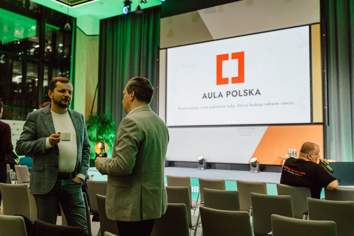 zdjecia eventowe aula polska bnp paribas warszawa 009 Dwóch mężczyzn stoi i rozmawia w sali konferencyjnej; jeden trzyma filiżankę. W tle duży ekran wyświetla logo i nazwę "AULA POLSKA". Widać także kilka pustych krzeseł i innego mężczyznę.