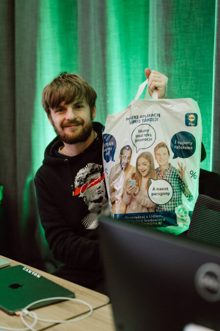 zdjecia eventowe aula polska bnp paribas warszawa 010 Uśmiechnięty mężczyzna z brodą siedzi przy biurku, trzymając białą torbę na zakupy z polskim tekstem i wizerunkami ludzi. Na biurku stoi laptop, a w tle wisi zielona zasłona.