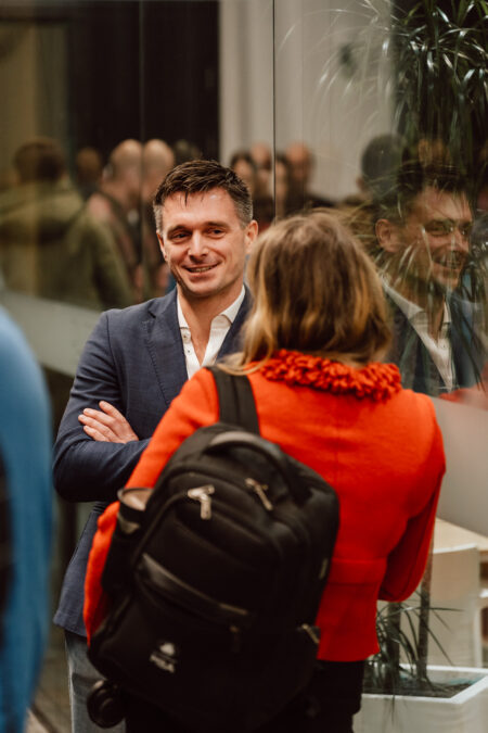 zdjecia eventowe aula polska bnp paribas warszawa 011 Mężczyzna w niebieskiej marynarce uśmiecha się ze skrzyżowanymi ramionami, rozmawiając z kobietą w czerwonej kurtce i czarnym plecaku. Znajdują się w pomieszczeniu, w pobliżu szklanej ściany z odbiciami i kilkoma roślinami.