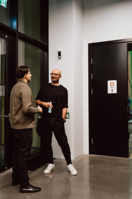 zdjecia eventowe aula polska bnp paribas warszawa 012 Dwóch mężczyzn stoi w korytarzu przy drzwiach oznaczonych "Aula", uśmiechając się i podając sobie ręce. Jeden z nich ma na sobie ciemny sweter i spodnie; drugi nosi okulary, czarny strój i białe trampki.