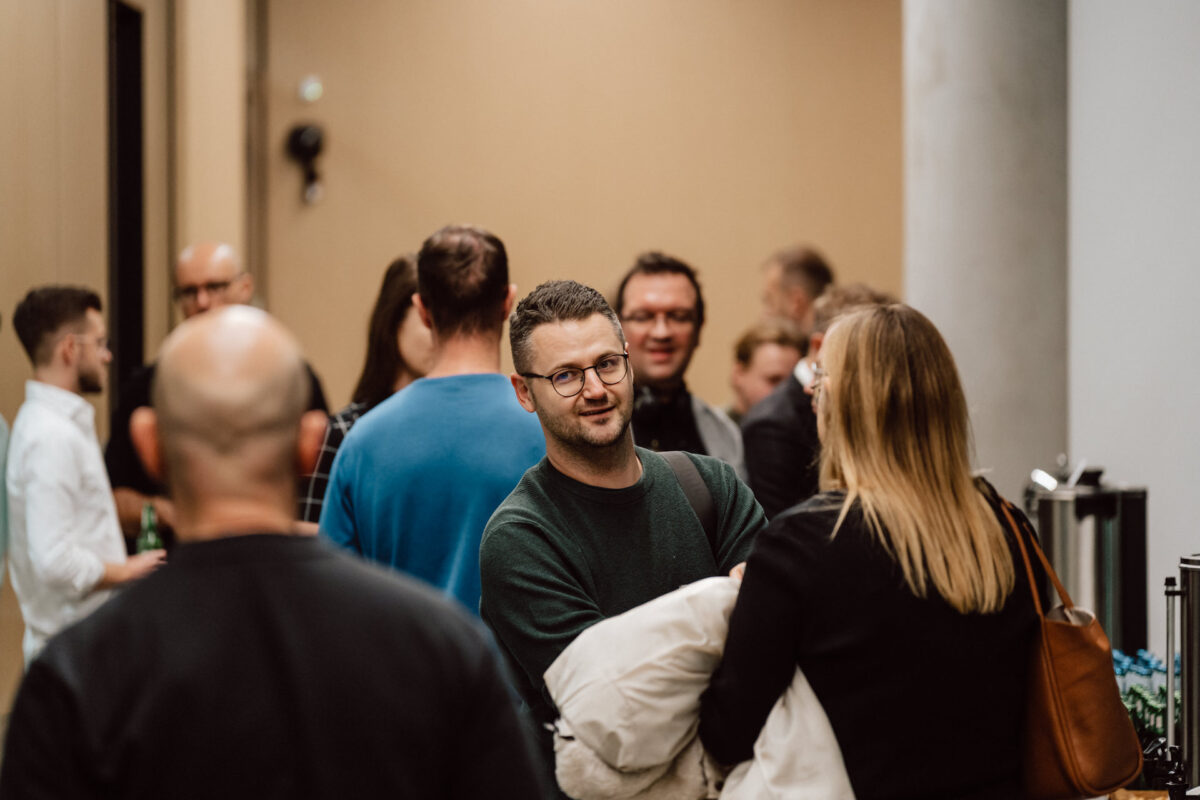 zdjecia eventowe aula polska bnp paribas warszawa 015 Grupa ludzi spotyka się w pomieszczeniu na czymś, co wydaje się być wydarzeniem networkingowym lub swobodnym spotkaniem. W centrum uwagi znajduje się mężczyzna w okularach i zielonym swetrze rozmawiający z kobietą o długich blond włosach.