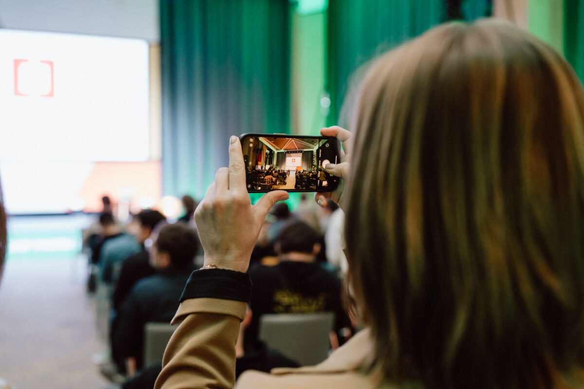 zdjecia eventowe aula polska bnp paribas warszawa 016 Osoba trzyma smartfon, aby zrobić zdjęcie lub nagrać film z prezentacji lub wydarzenia w dużym pomieszczeniu wypełnionym siedzącymi uczestnikami, z zielonymi zasłonami i jasnym ekranem w tle.