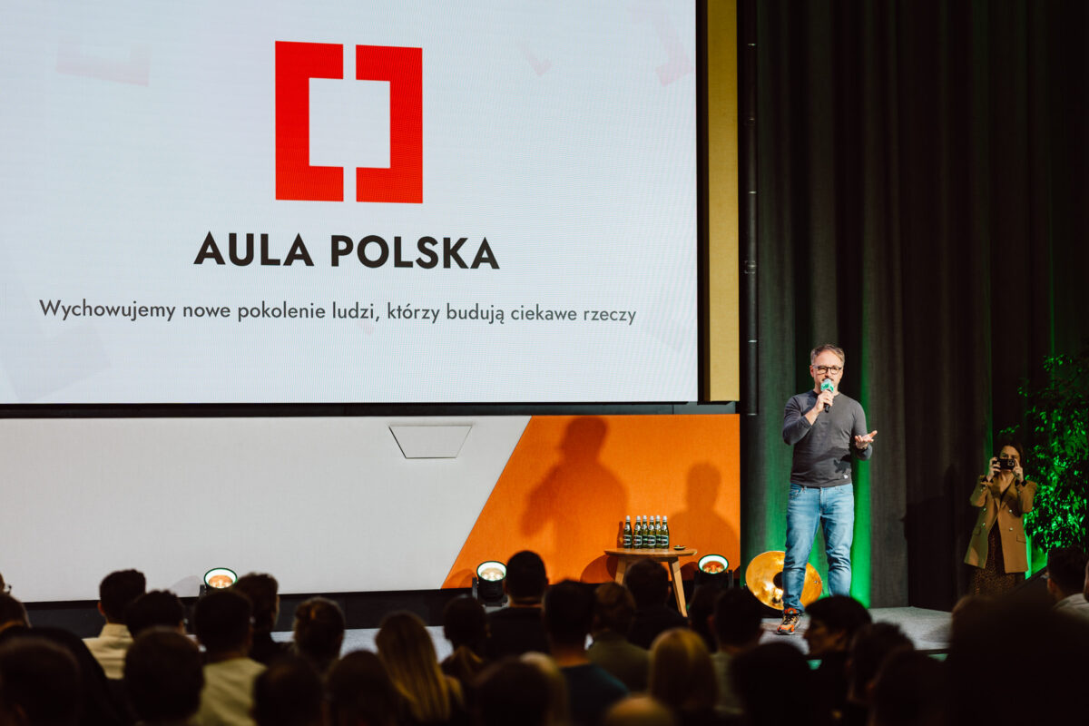 zdjecia eventowe aula polska bnp paribas warszawa 018 Mężczyzna przemawia na scenie z mikrofonem podczas wydarzenia Aula Polska. Za nim na ekranie wyświetlane jest logo Aula Polska i polski tekst. Publiczność patrzy, a w pobliżu stoi fotograf.