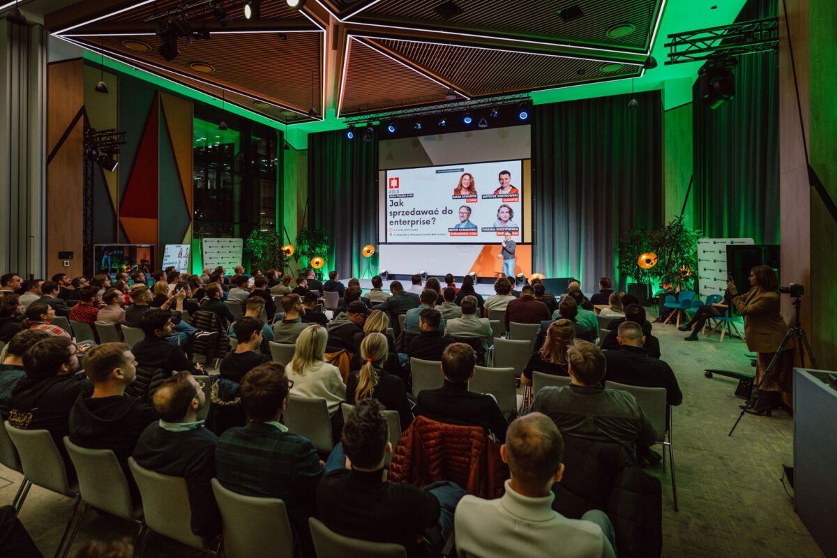 zdjecia eventowe aula polska bnp paribas warszawa 019 Duża publiczność siedzi naprzeciwko sceny z panelem prelegentów i dużym ekranem wyświetlającym ich zdjęcia i temat dyskusji w nowoczesnej, dobrze oświetlonej przestrzeni eventowej z zielonymi akcentami świetlnymi.