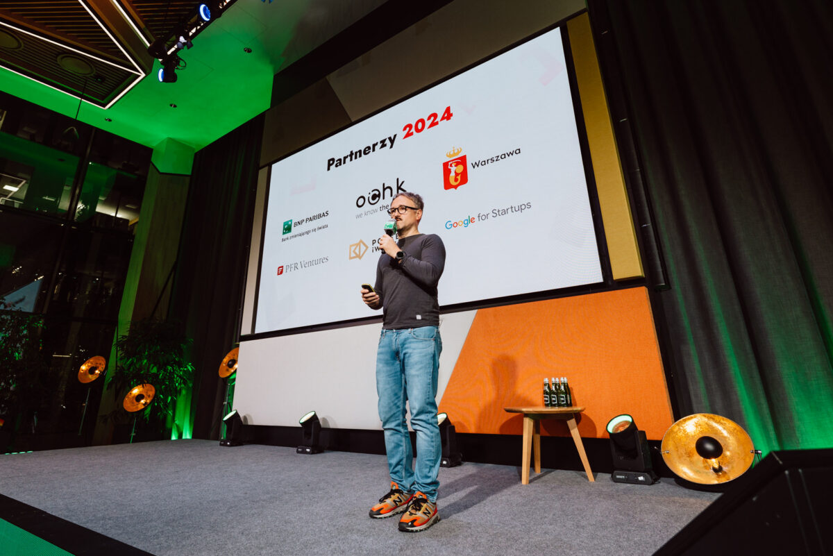 zdjecia eventowe aula polska bnp paribas warszawa 020 Osoba w dżinsach i swetrze przemawia na scenie z mikrofonem. Za nimi na ekranie wyświetlany jest napis "Partners 2024" i logotypy różnych organizacji. Wydaje się, że jest to nowoczesne, kryte miejsce wydarzeń.