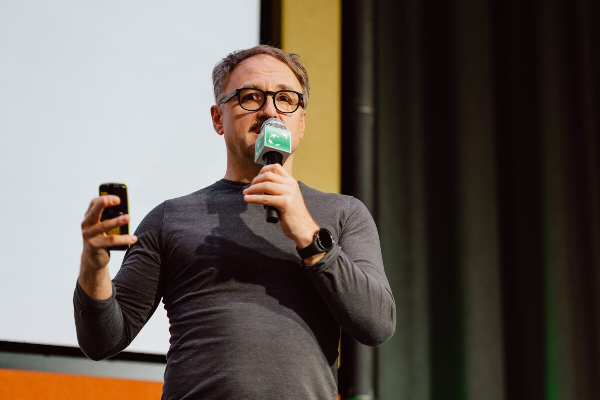 zdjecia eventowe aula polska bnp paribas warszawa 021 Mężczyzna w okularach i szarej koszuli z długim rękawem mówi do mikrofonu, trzymając smartfon, stojąc przed ekranem podczas prezentacji.