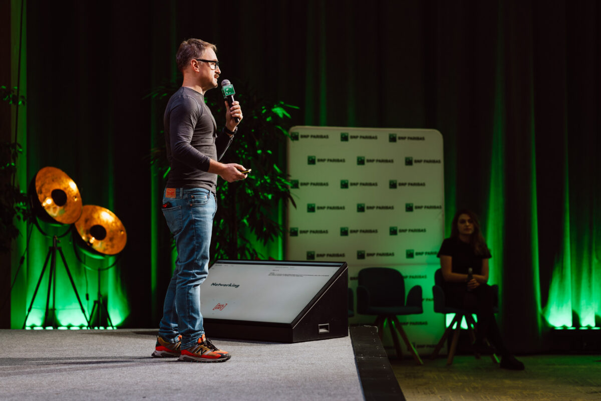 zdjecia eventowe aula polska bnp paribas warszawa 022 Mężczyzna w dżinsach i ciemnej koszuli mówi do mikrofonu na scenie z zielonym oświetleniem, reflektorami i tłem marki BNP Paribas; członek publiczności siedzi w pobliżu.