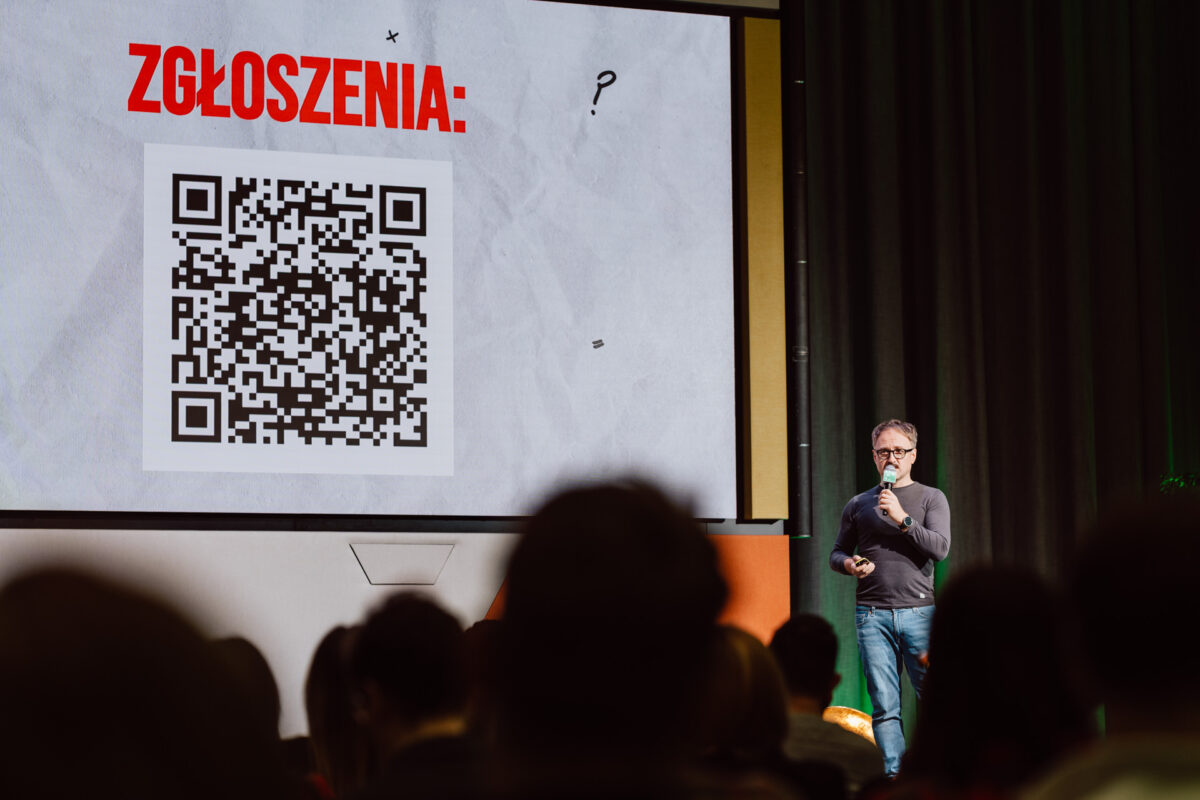 zdjecia eventowe aula polska bnp paribas warszawa 024 Na scenie stoi mężczyzna mówiący do mikrofonu przed ekranem wyświetlającym duży kod QR i słowo "ZGŁOSZENIA". Członkowie publiczności są widoczni na pierwszym planie.