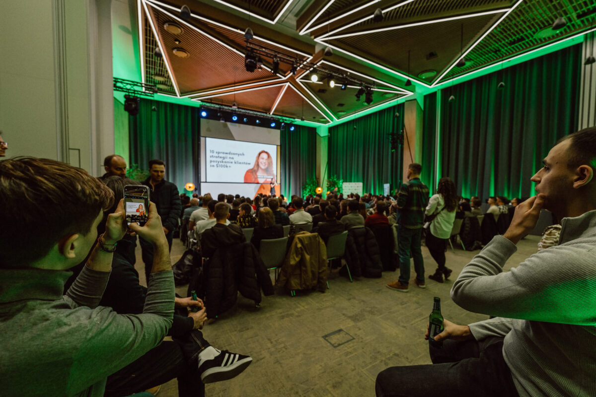 zdjecia eventowe aula polska bnp paribas warszawa 025 Duża publiczność siedzi w nowoczesnej, oświetlonej na zielono sali konferencyjnej, obserwując kobietę prezentującą na ekranie z przodu. Niektórzy uczestnicy robią zdjęcia i trzymają napoje. Sufit zdobi geometryczne oświetlenie.