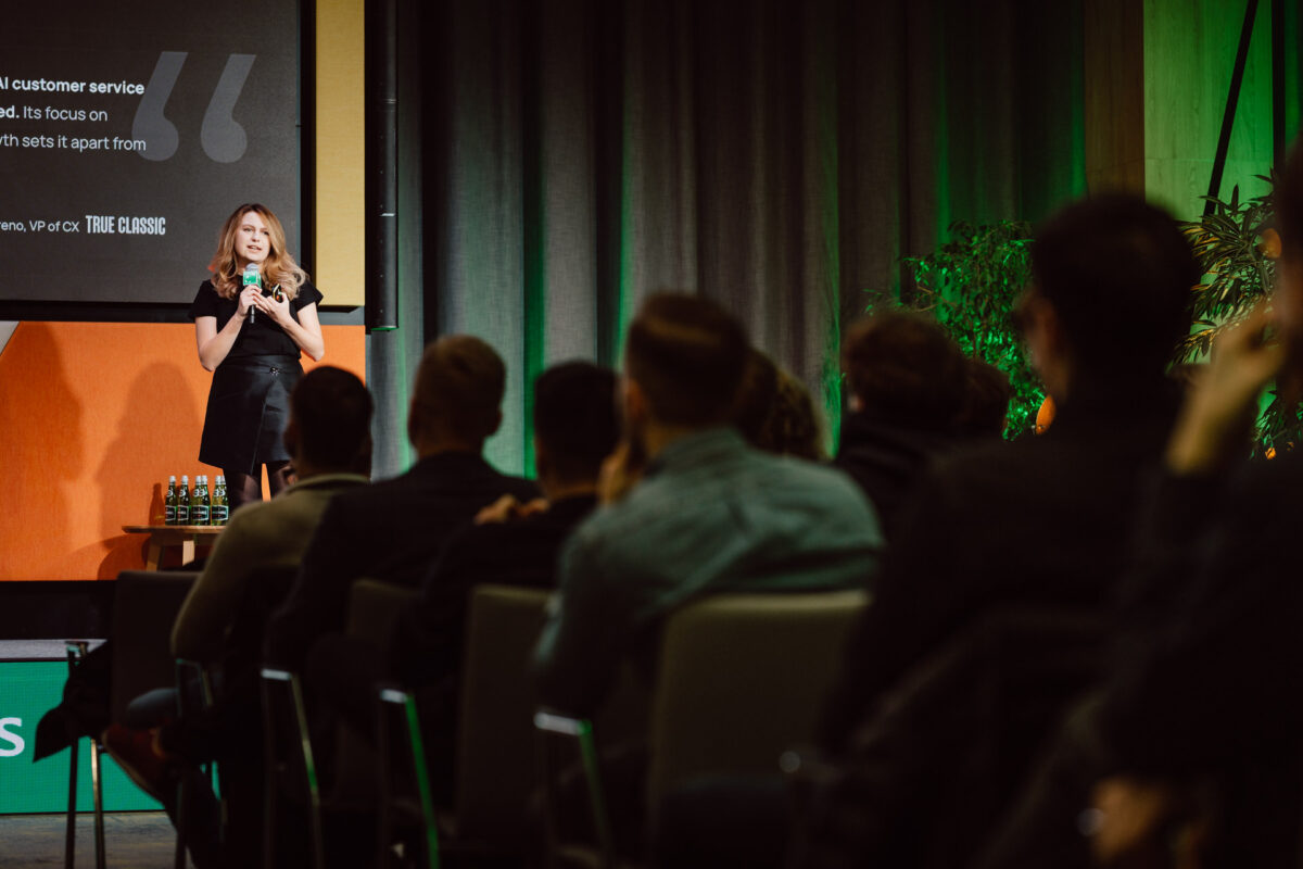 zdjecia eventowe aula polska bnp paribas warszawa 026 Kobieta w czarnej sukience stoi na scenie, przemawiając do publiczności siedzącej w rzędach. Za nią znajduje się cytat wyświetlany na ekranie i zielone rośliny z boku. Na stole obok sceny widoczne są butelki.